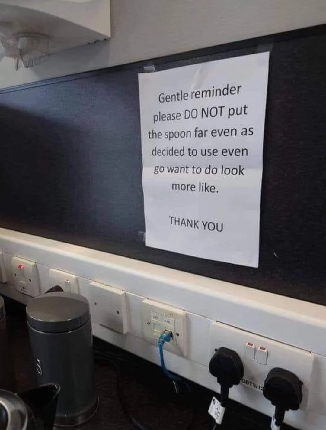 A white paper sign taped to a dark wall above a row of electrical outlets and sockets, with a small trash can visible below…