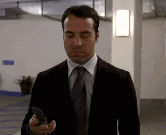 A man in a suit walks through a parking garage, looking down at his phone, then turns and gestures dismissively at a potted…