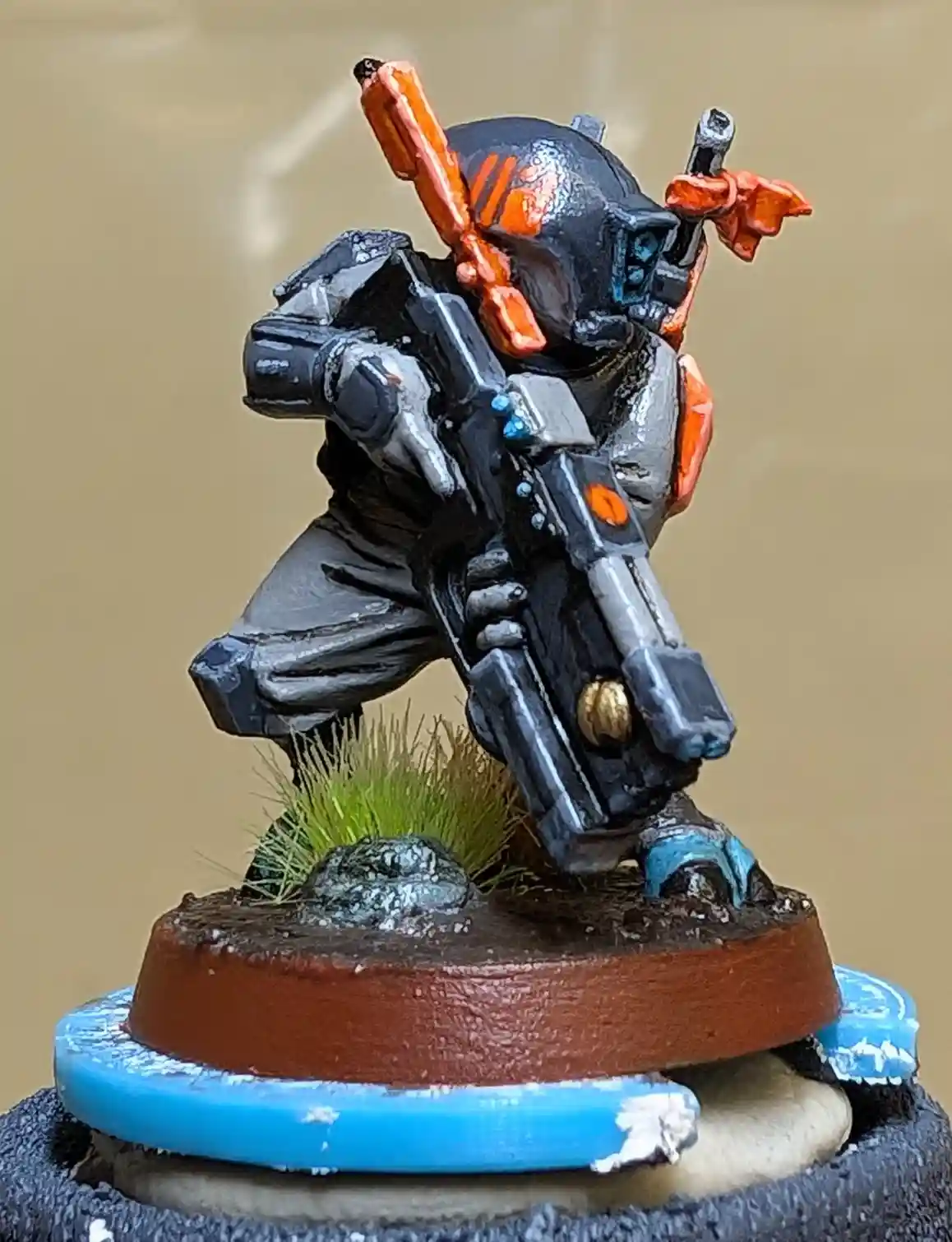 A painted miniature of a futuristic soldier in black and orange armor, holding a large gun, stands on a base with green…