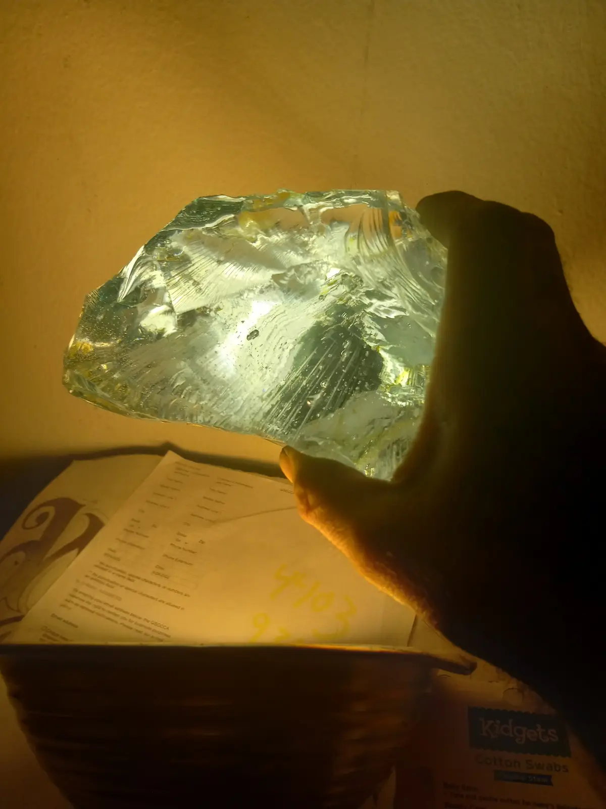 A hand holds a large, irregularly shaped, translucent ice chunk with visible internal fractures and air bubbles, illuminated…