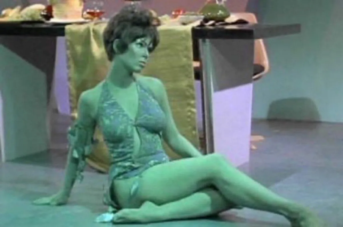 A woman with green-tinted skin sits on the floor in a patterned halter top, looking displeased. The image has a green color…