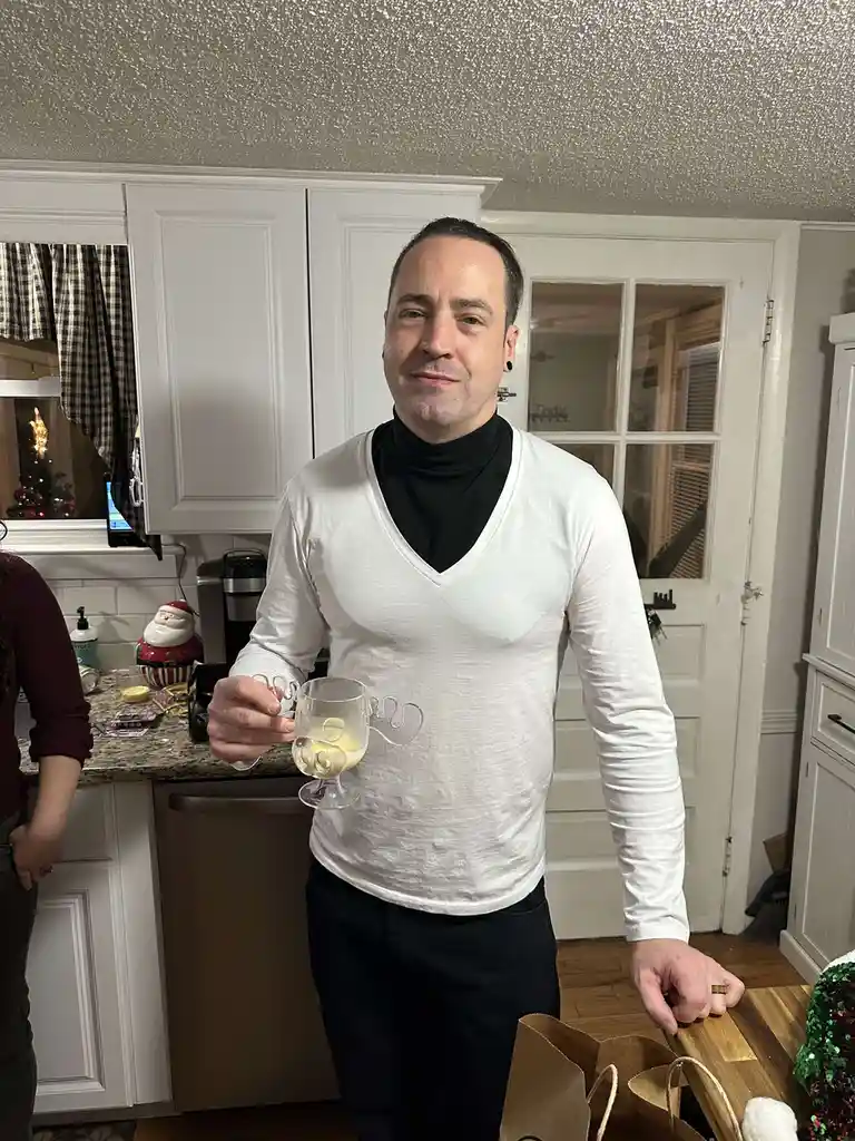 A man in a white v-neck shirt over a black turtleneck stands in a kitchen holding a glass of champagne, smiling slightly…