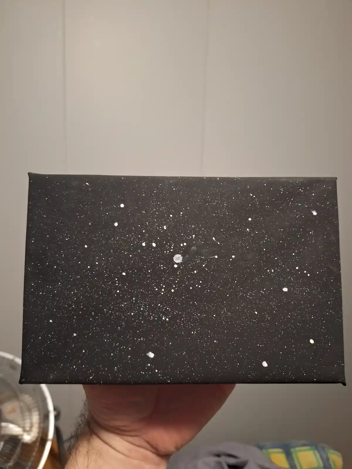 A hand holds up a rectangular black canvas painted with numerous white dots resembling stars, with a few larger, smudged…