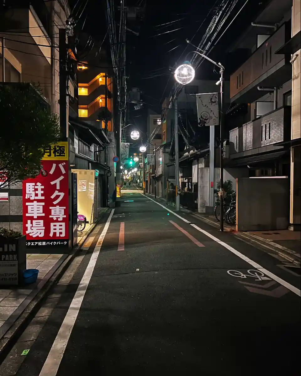 A nighttime photo of a quiet, narrow Japanese street with illuminated spherical streetlights and buildings on both sides. A…