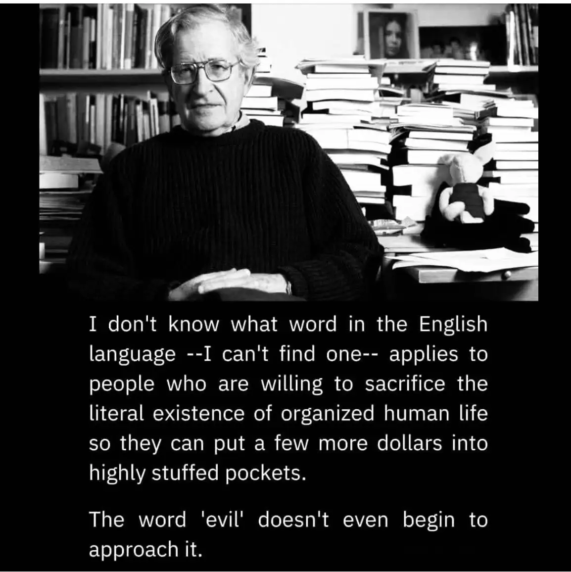 A black-and-white photo of Noam Chomsky sitting at a desk surrounded by stacks of books. He wears glasses and a dark…