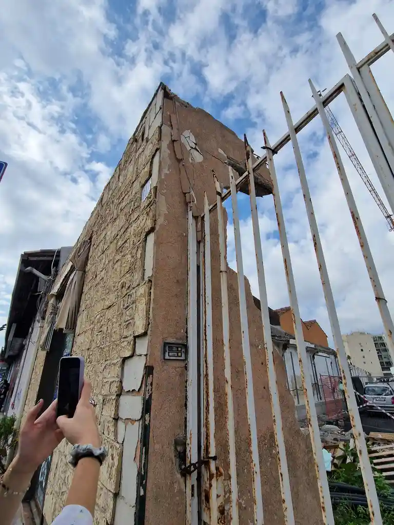 A person holds a smartphone to photograph a dilapidated, partially demolished building with exposed stone and crumbling…