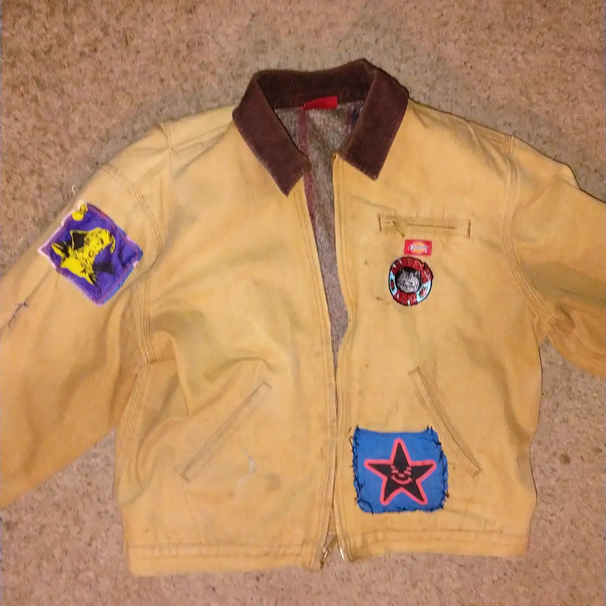 A yellow jacket with a brown collar lies flat on a beige carpet, covered in multiple colorful patches including a purple one…