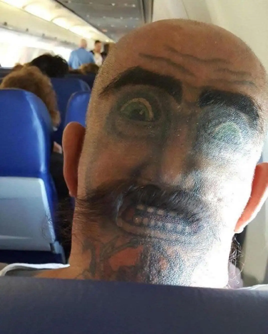 A bald man is seated on an airplane, his entire face and neck covered in a detailed, cartoonish tattoo depicting a…