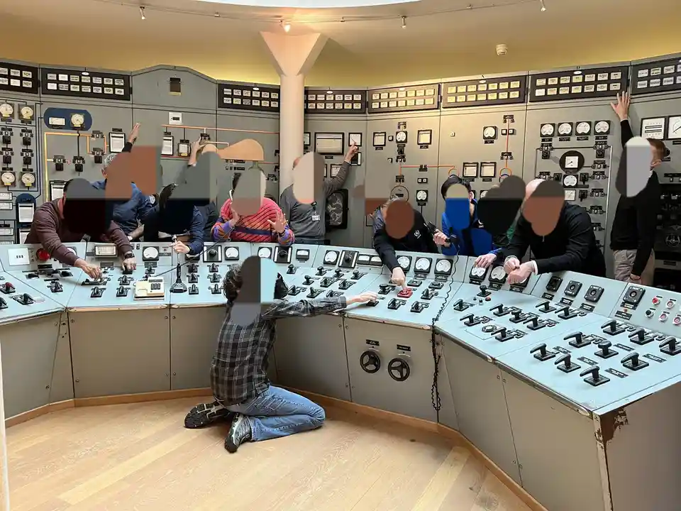 A group of people are gathered around a large, vintage control room console filled with dials, switches, and gauges. Most…