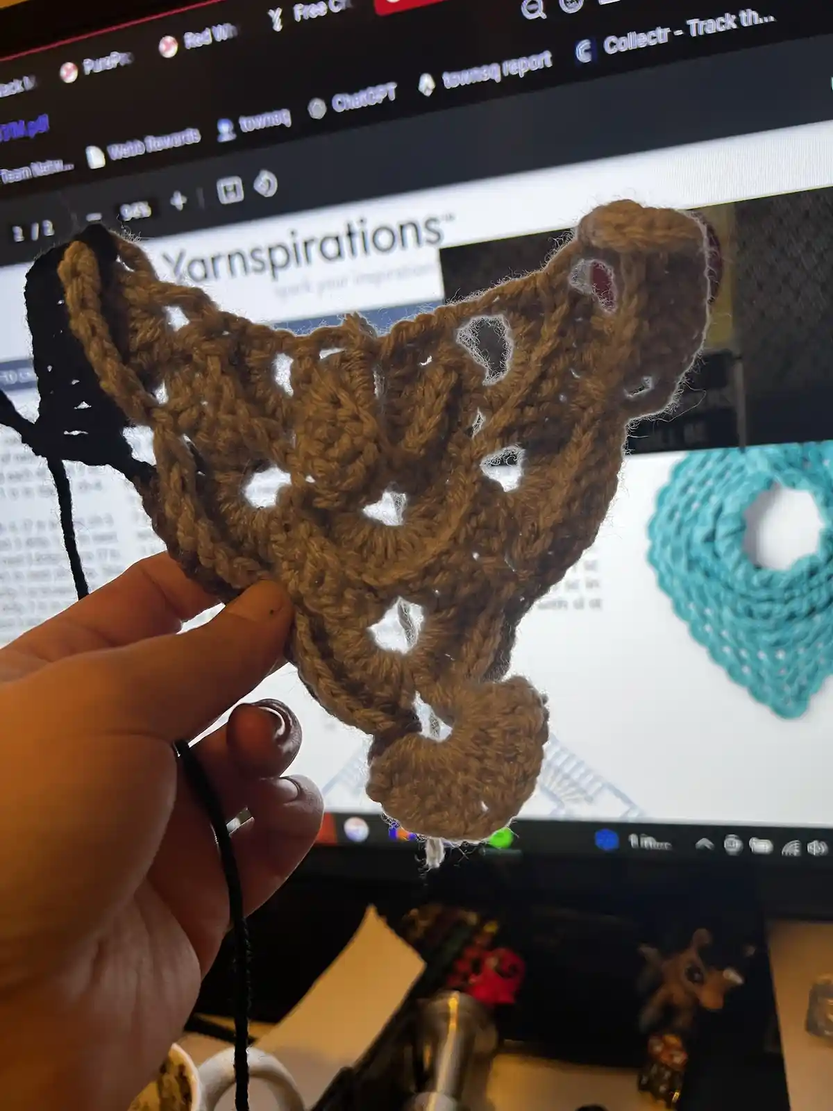 A hand holds up a brown, crocheted piece shaped like a butterfly, which is visibly lopsided and misshapen compared to the…