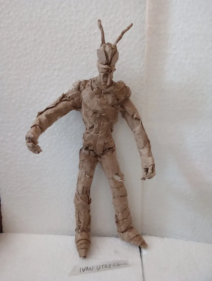 A handmade figure crafted from brown paper or cardboard, standing upright with arms slightly outstretched and two twig-like…