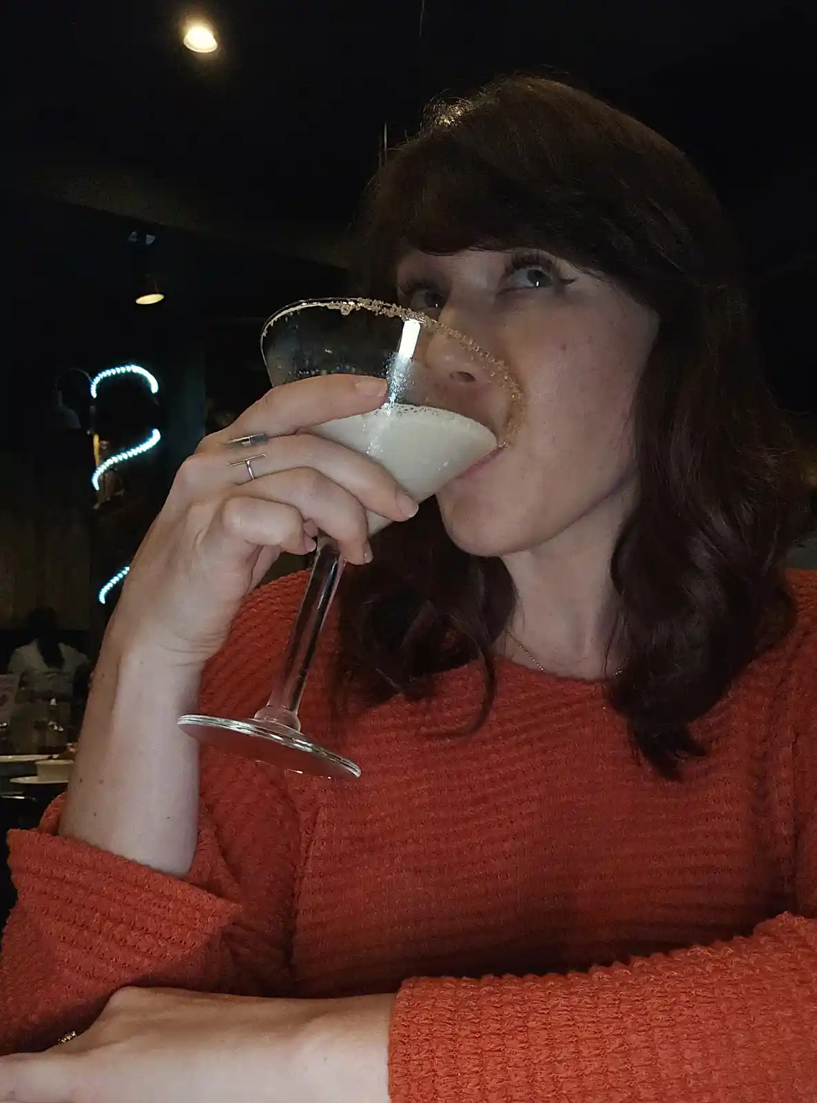A woman in an orange sweater is drinking from a cocktail glass with a sugared rim, looking upward with a skeptical…
