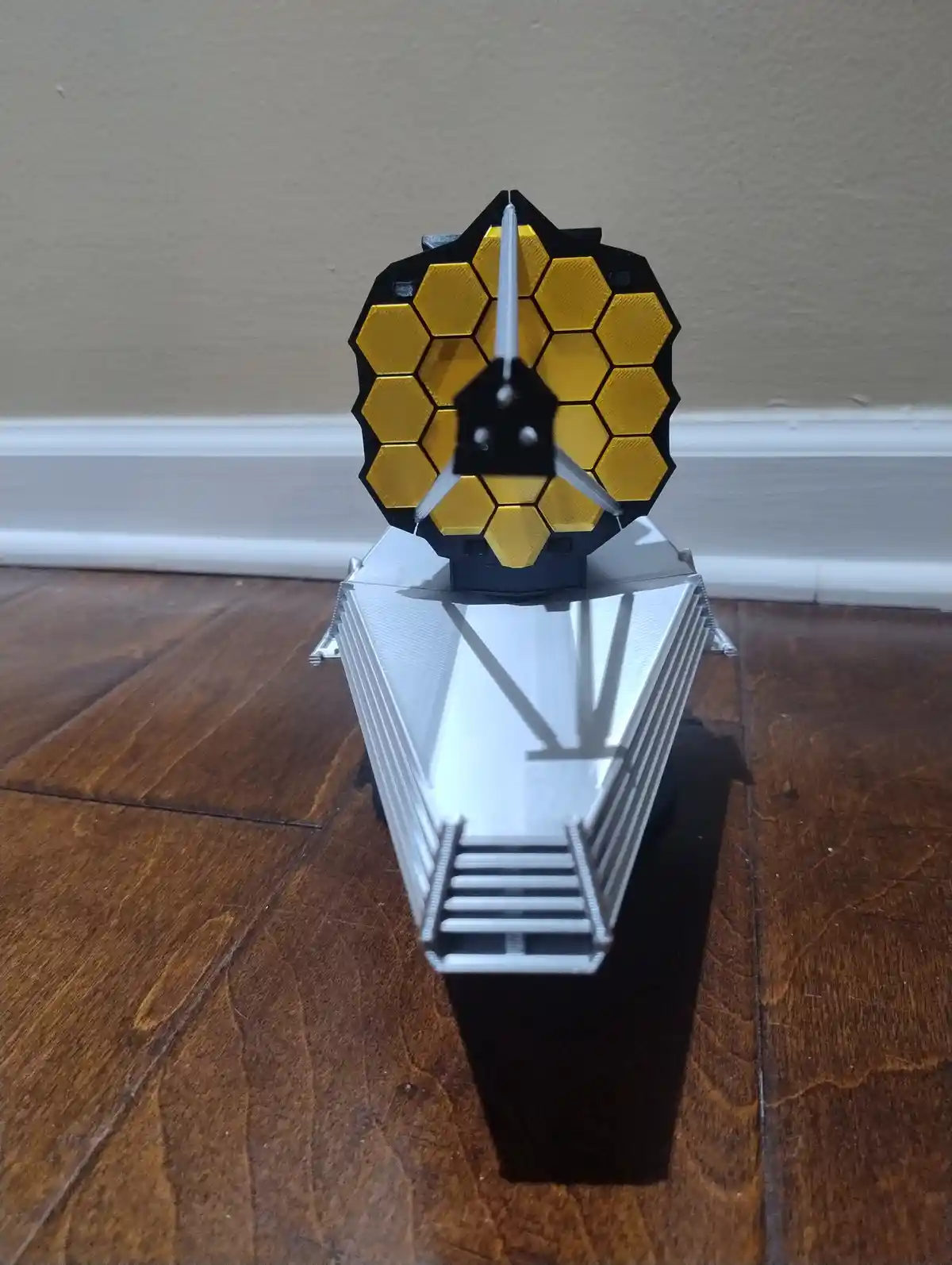A 3D-printed model of the James Webb Space Telescope sits on a wooden floor, its hexagonal gold mirror array and silver…