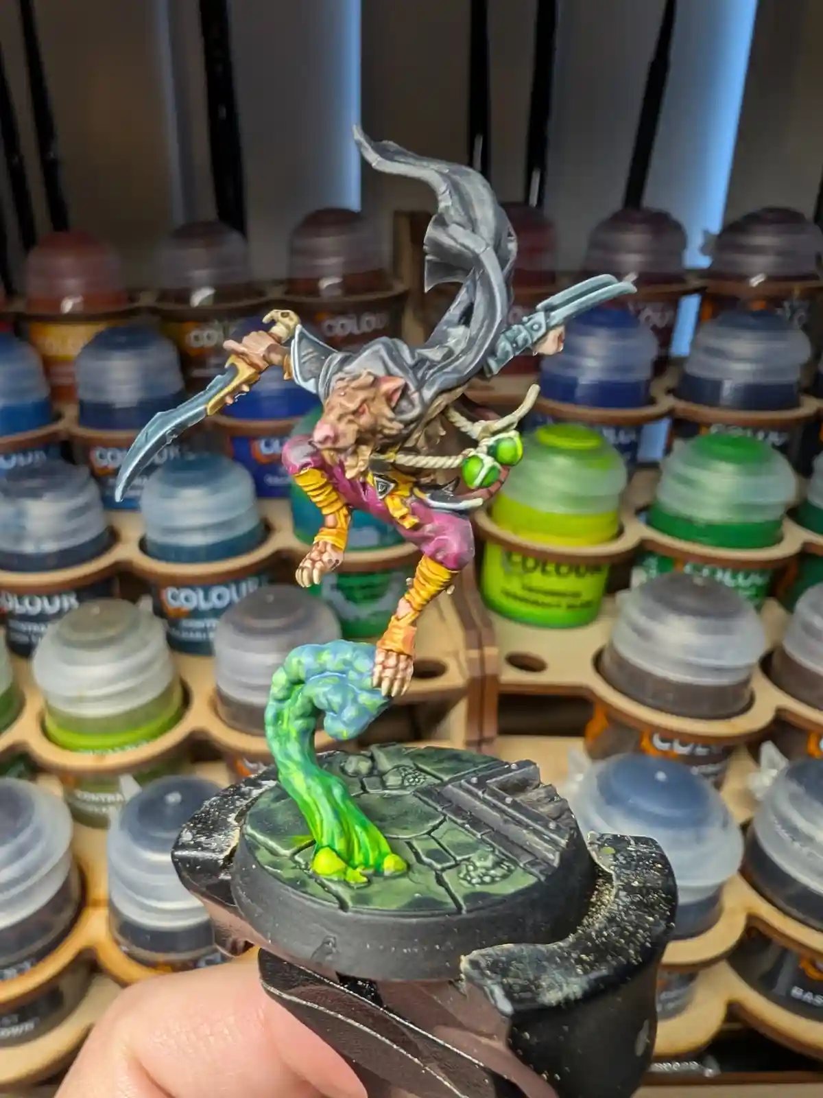 A hand holds a painted miniature of a rat-like humanoid warrior with a flowing grey cape, wielding two curved swords…