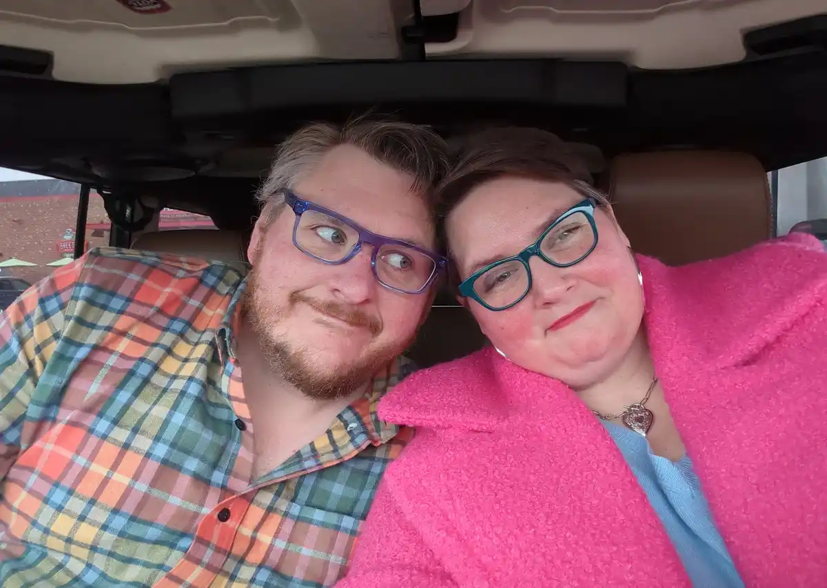 A selfie of a man and woman sitting in the front seats of a car, both wearing glasses and looking at the camera. The man on…