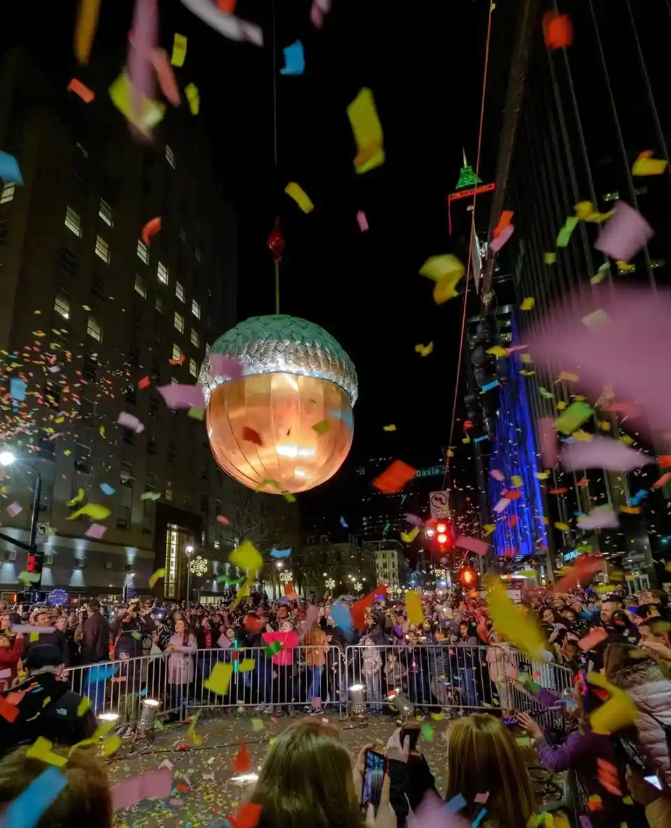A nighttime street scene shows a large, glowing, spherical object resembling a giant acorn or disco ball suspended above a…