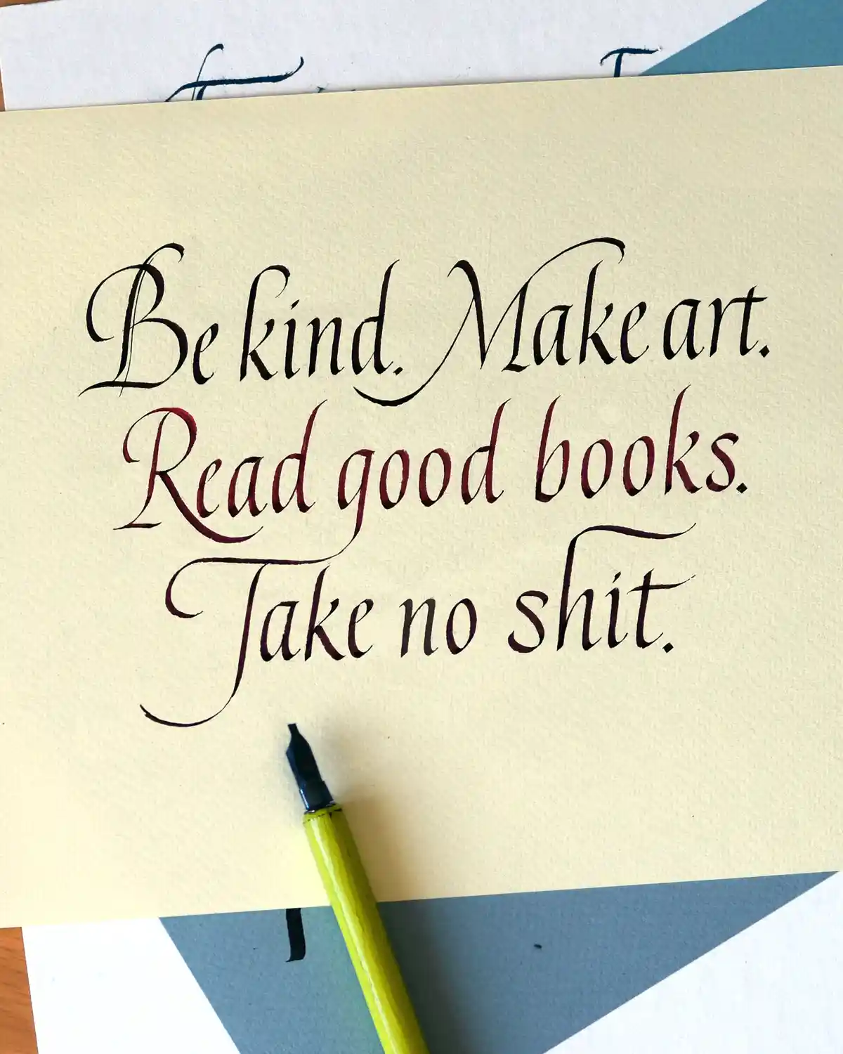 A close-up of elegant calligraphy on cream-colored paper, written with a fountain pen. The text reads 'Be kind. Make art…
