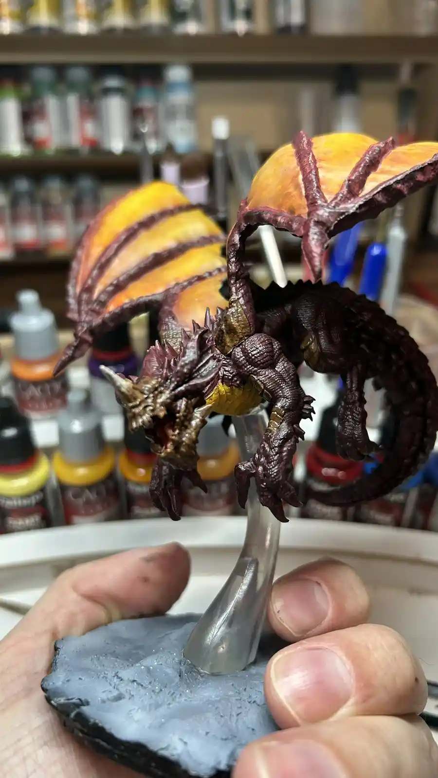 A hand holds a detailed, painted miniature dragon figurine with large orange and yellow wings and dark purple scales…