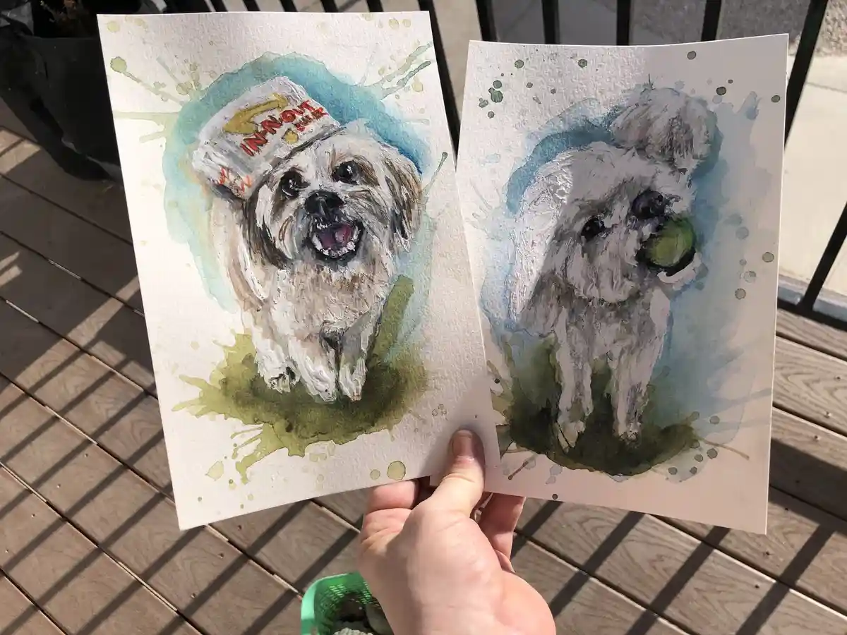 A hand holds two watercolor dog portraits side by side; the left shows a white dog wearing an 'In-N-Out Burger' hat, and the…
