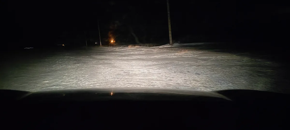 A nighttime photo taken from inside a car, showing headlights illuminating a snowy, icy road ahead. The scene is dark with…