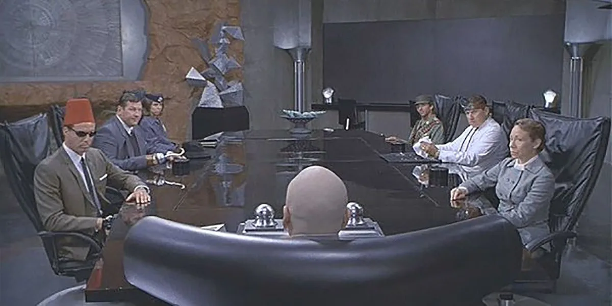 A still from the movie 'The World Is Not Enough' shows a tense meeting around a large black table. A bald man sits at the…