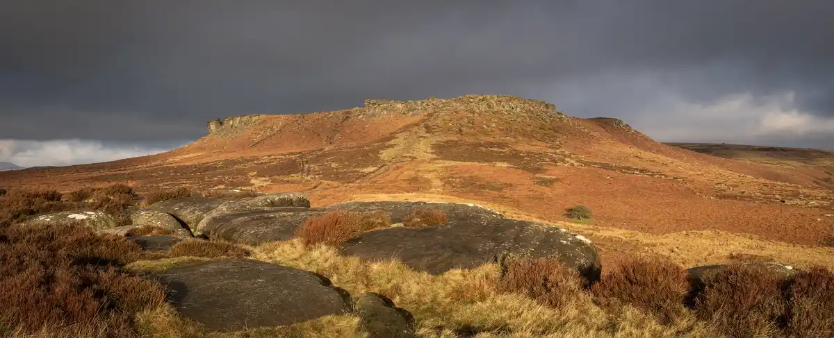 A wide landscape photo shows a rugged, rocky hill under a dark, cloudy sky, with reddish-brown heather and grass covering…