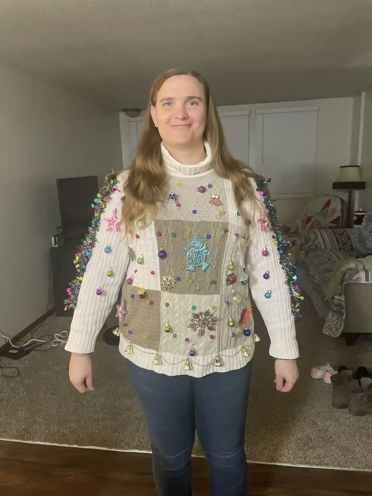 A woman stands indoors wearing a heavily decorated, multi-colored Christmas sweater adorned with numerous ornaments, tinsel…