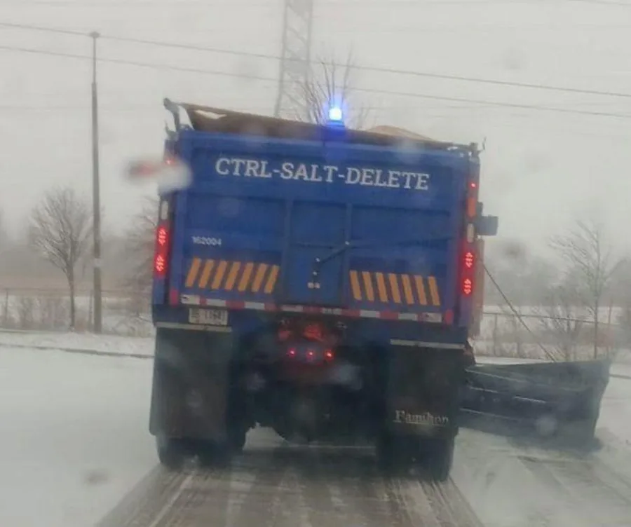 A blue snowplow truck is seen from behind on a snowy road, its rear has the text 'CTRL-SALT-DELETE' painted on it, with a…