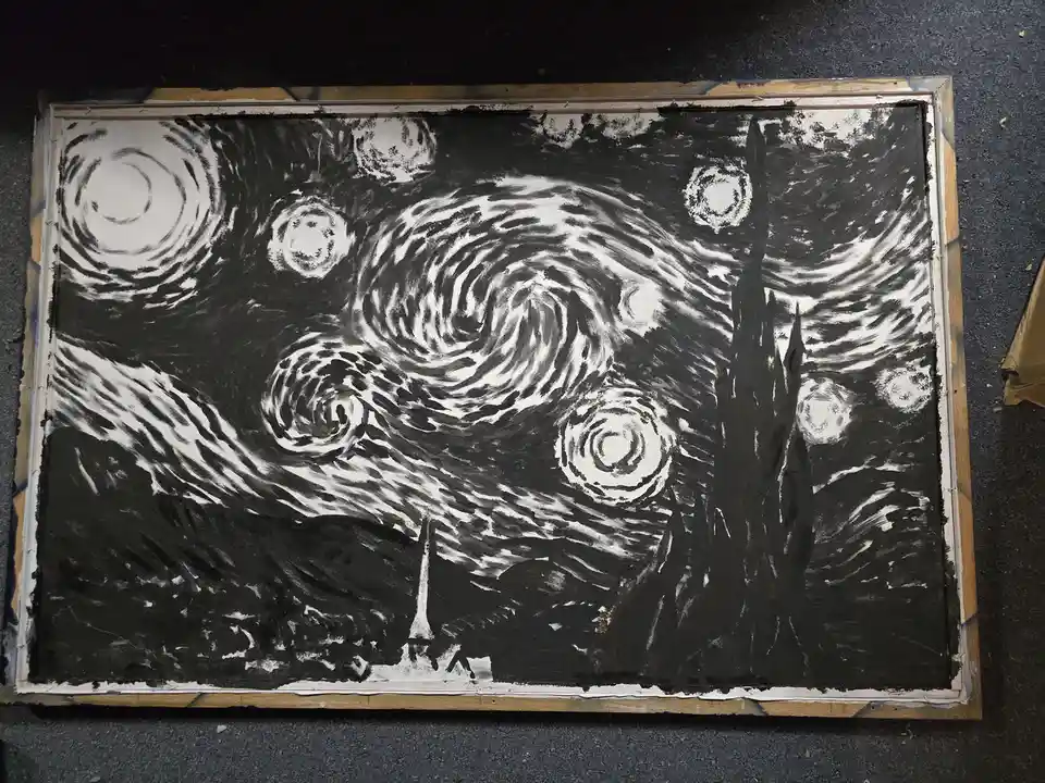 A black-and-white painting of Vincent van Gogh's 'Starry Night' is shown in a wooden frame, resting on a dark floor. The…