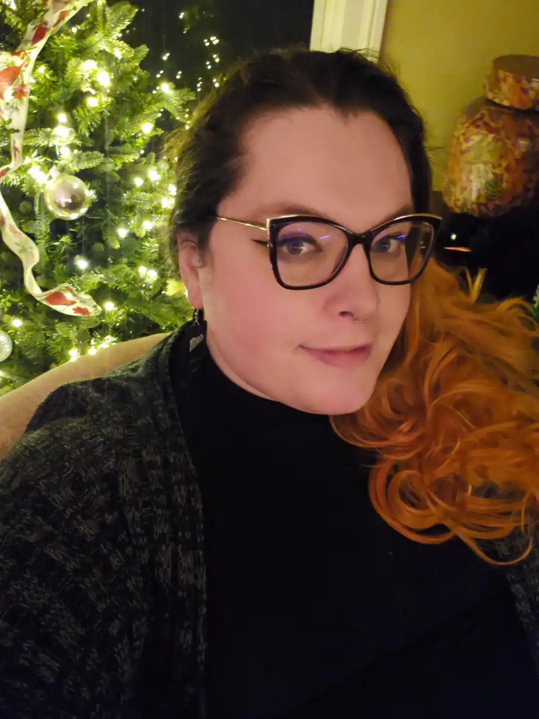 A person with glasses and reddish hair sits in front of a lit christmas tree, wearing a black turtleneck and a dark…