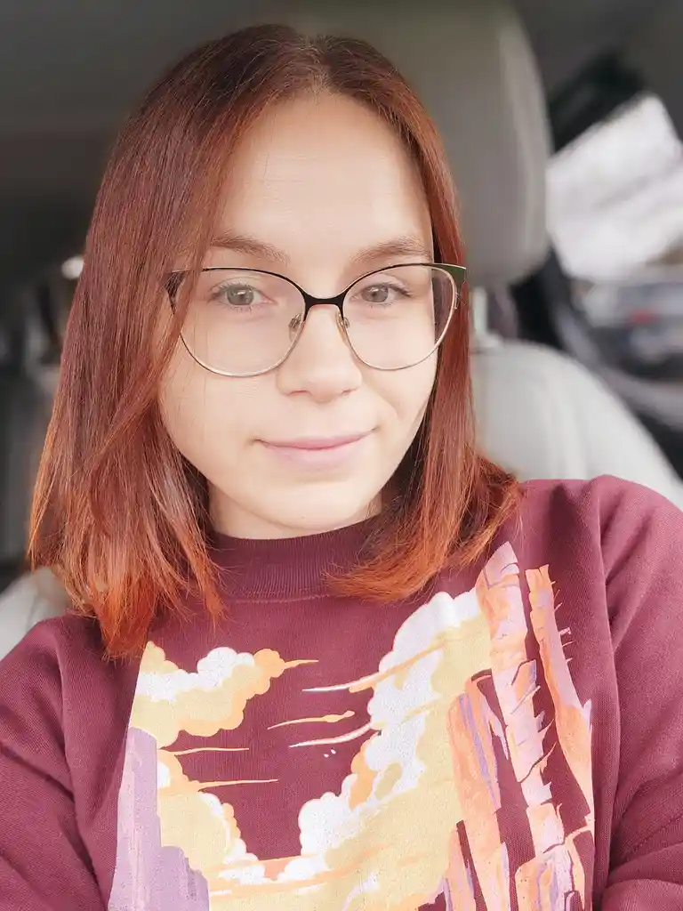 A person with reddish-brown hair and glasses, wearing a maroon sweatshirt with a canyon/cloud graphic, sits in a car and…
