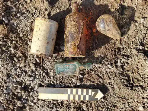 An overhead shot shows four weathered, dirt-covered bottles and a ceramic-like cylinder lying on rough, gravelly soil. A…