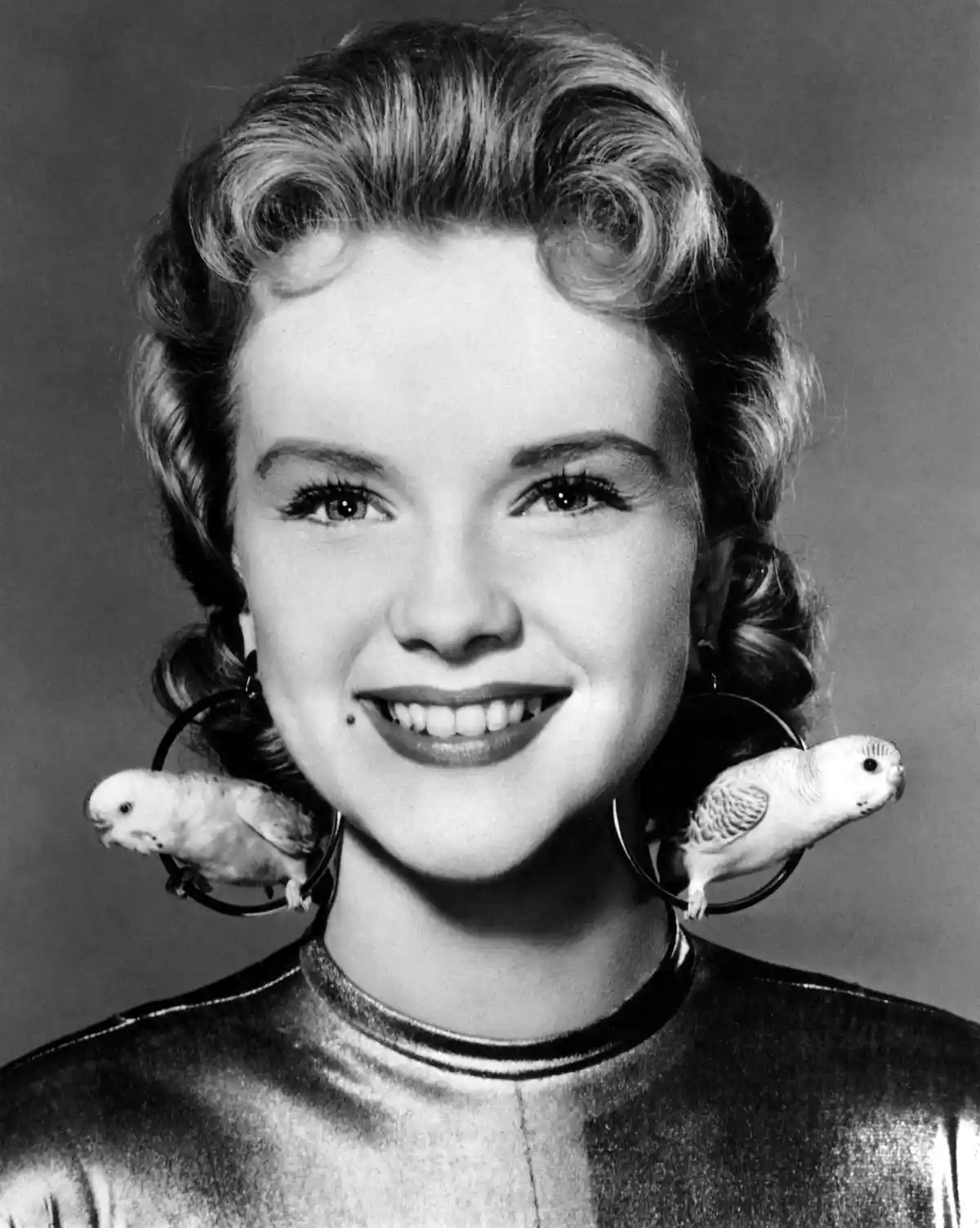 A black-and-white portrait of a smiling woman with vintage curled hair and large hoop earrings, each holding a small white…