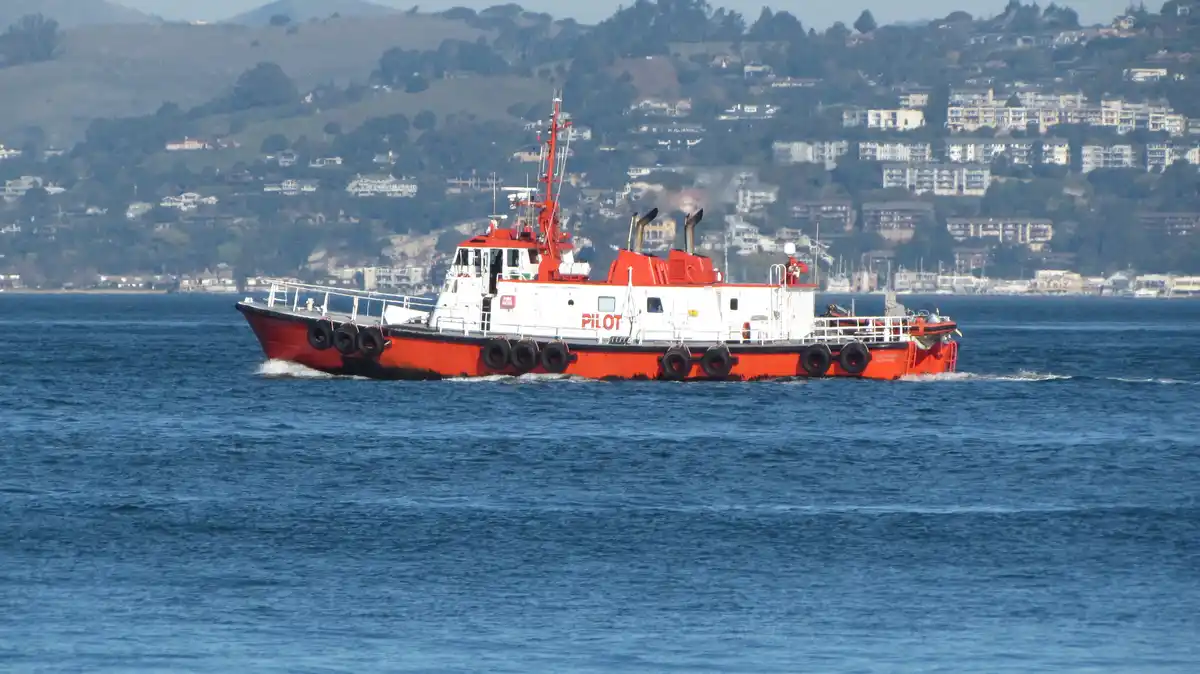 A red and white pilot boat with 'PILOT' written on its side is moving through blue water, leaving a wake. In the background…