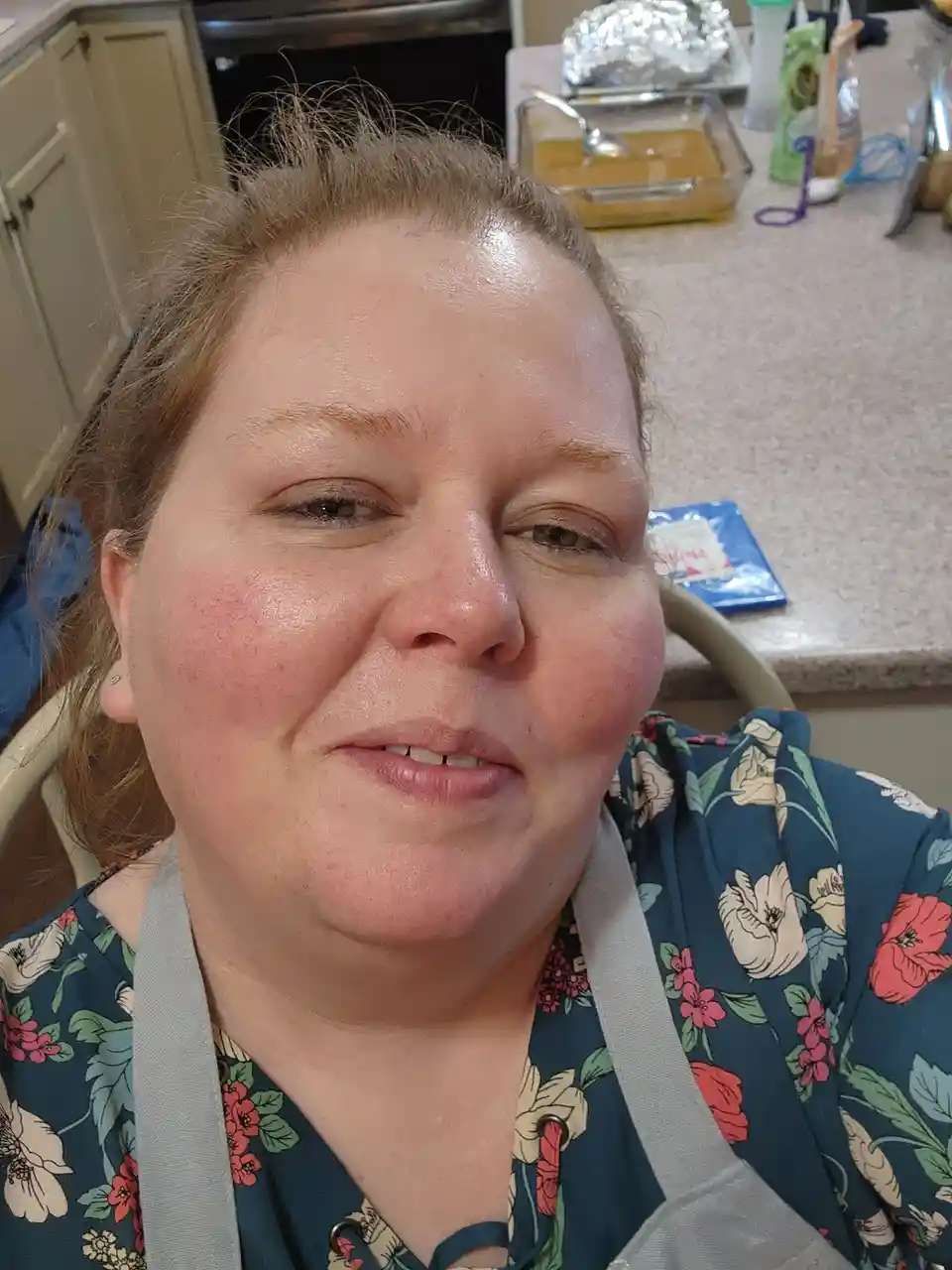 A woman with flushed cheeks and a slightly tired expression sits in a kitchen, wearing a floral shirt and an apron. Behind…