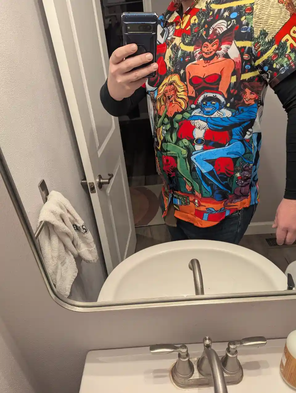 A person takes a mirror selfie in a bathroom, wearing a very colorful and busy Christmas-themed shirt featuring comic…