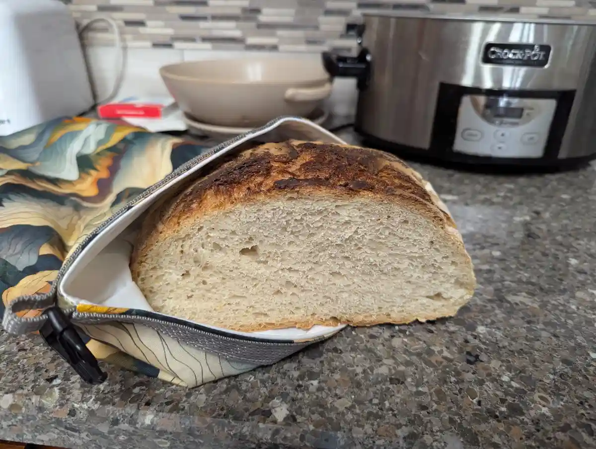 A loaf of artisan bread with a dark, unevenly browned crust sits partially inside an open, colorful zippered fabric bag on a…