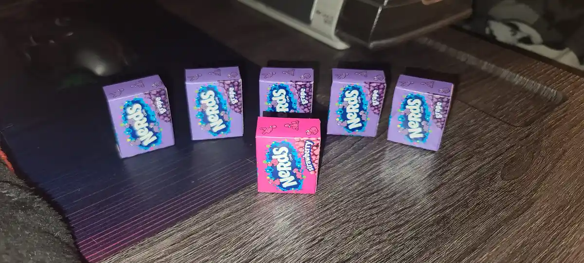 Six small boxes of Nerds candy are arranged on a dark wooden surface, five purple grape-flavored boxes and one pink…
