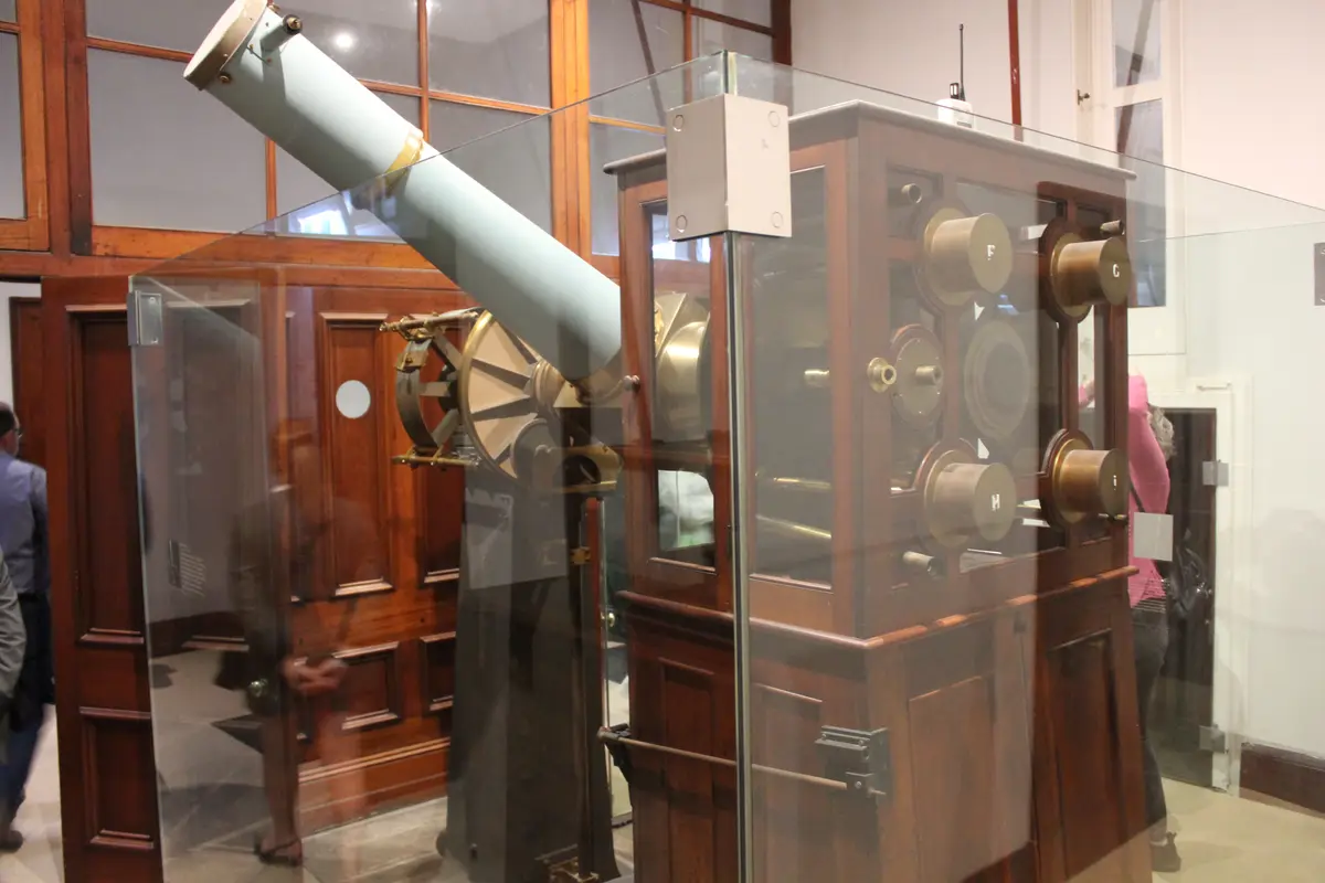 An old-fashioned, wooden-framed telescope with brass fittings is displayed behind glass in a museum, its barrel angled…