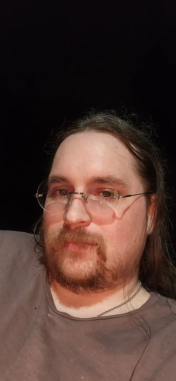 A close-up selfie of a man with long brown hair, a mustache and goatee, wearing round glasses and a beaded necklace, looking…