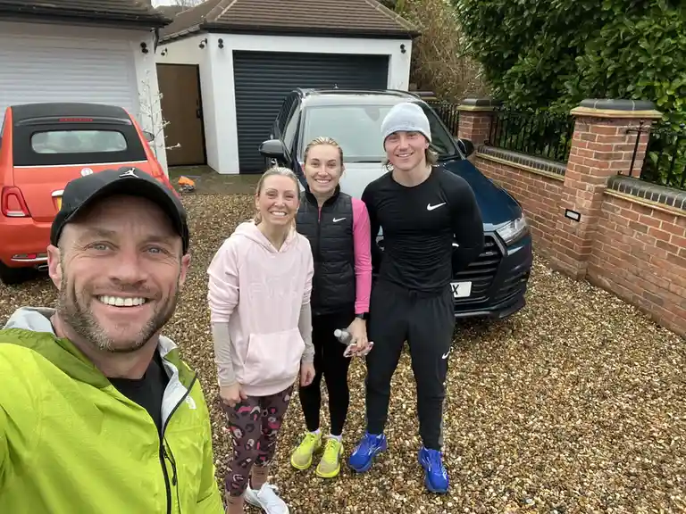 A selfie of four people in athletic wear standing on a gravel driveway in front of two parked cars, smiling at the camera…