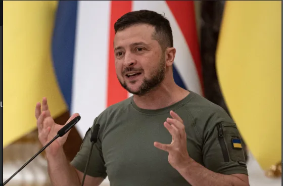 a man with a beard and short dark hair, wearing a green t-shirt with a small Ukrainian flag patch on the sleeve, stands in…