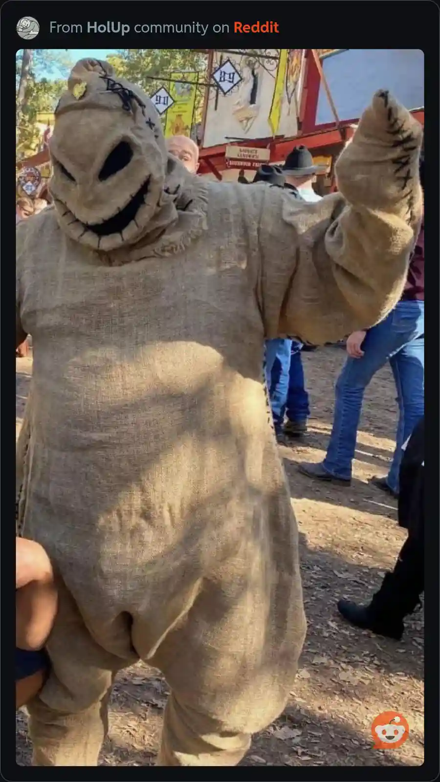 A person in a burlap scarecrow costume with a stitched smile and black eye holes stands outdoors, one arm raised. The…
