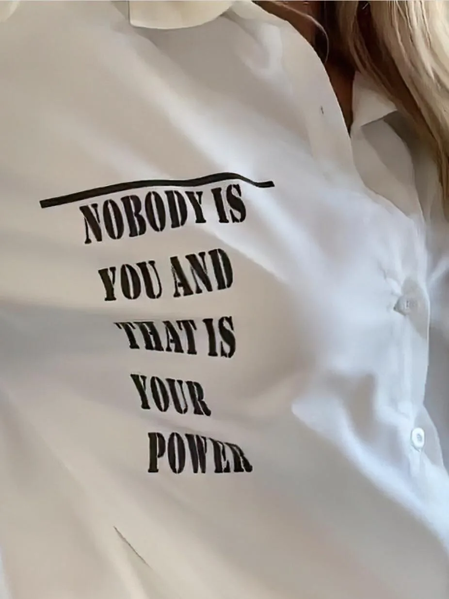 A close-up photo shows a person wearing a white button-up shirt with bold black text printed on the chest that reads 'NOBODY…