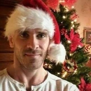A man with a neutral to slightly smug expression wears a red and white Santa hat while standing in front of a decorated…