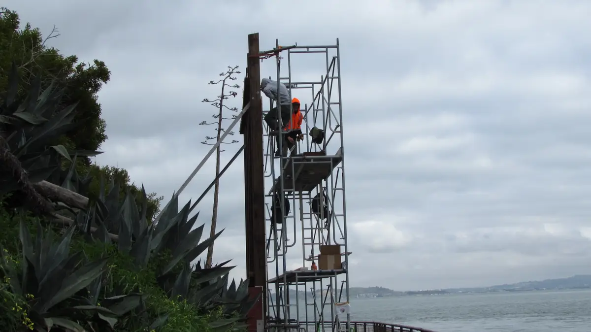 Two workers are on a tall metal scaffolding structure near a body of water, one in an orange safety vest and hard hat, the…