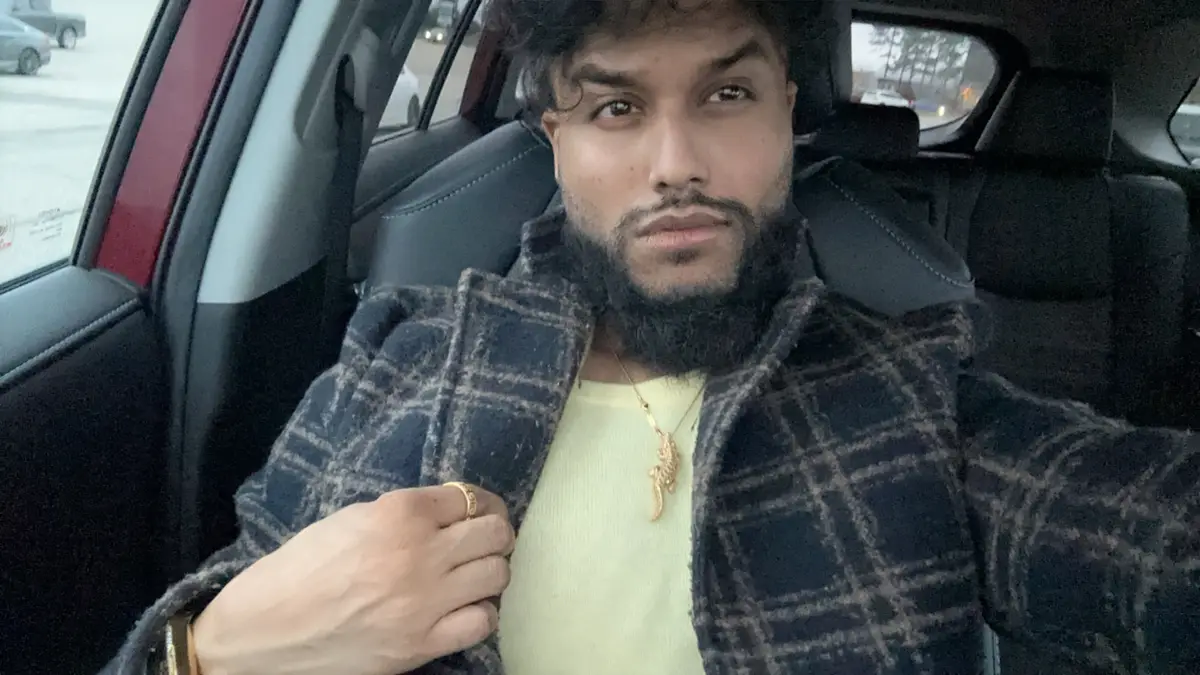 A man with a beard and dark curly hair sits in the driver's seat of a car, wearing a plaid jacket over a light yellow shirt…