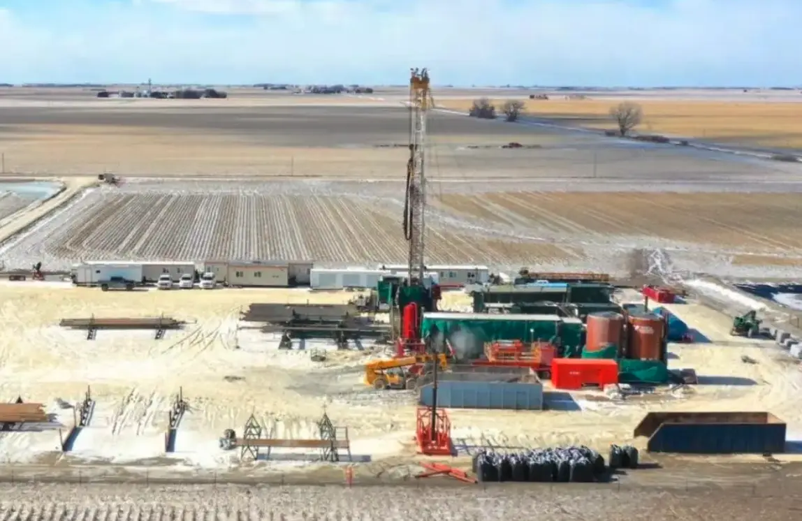 An aerial view of a large oil drilling rig in a flat, snowy, rural landscape with fields and temporary buildings. The…