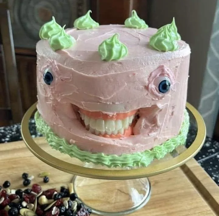 a round pink-frosted cake decorated to resemble a smiling monster with large blue eyes, a wide grin showing white teeth, and…