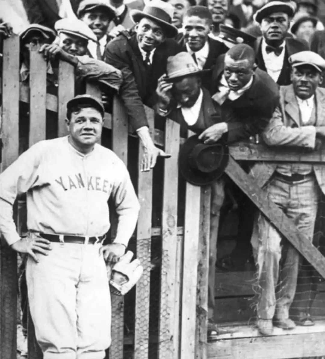 A black-and-white historical photo shows a baseball player in a 'YANKEE' uniform standing with hands on hips, while a crowd…